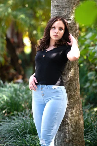 Young woman in a black top and jeans standing beside a tree